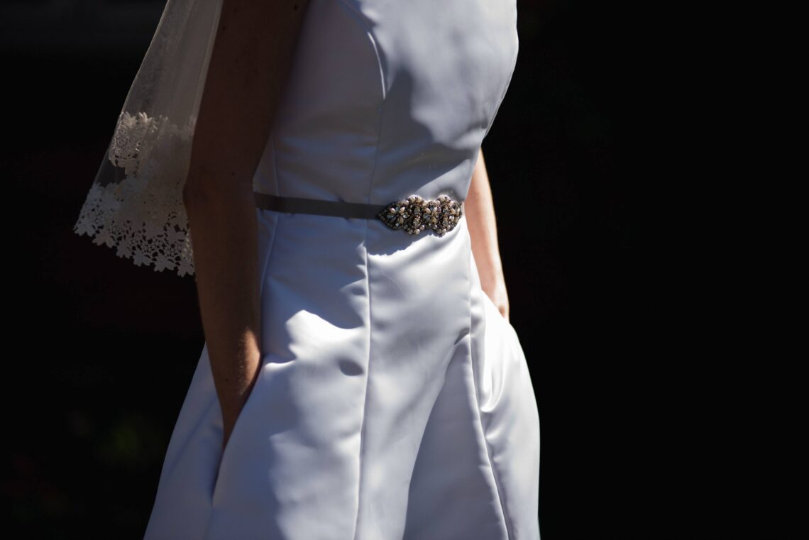Detail photo of Amy wearing her wedding dress; her face is not showing and her hands are in the pockets. The bottom of her veil is visible.