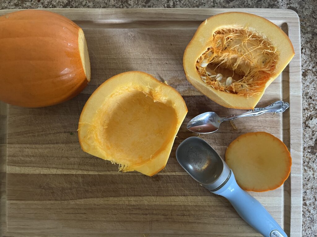 Two pie pumpkins; one is sliced in half, and one of the halves has had its flesh and seeds scraped out. An ice cream scoop and grapefruit spoon are also visible.