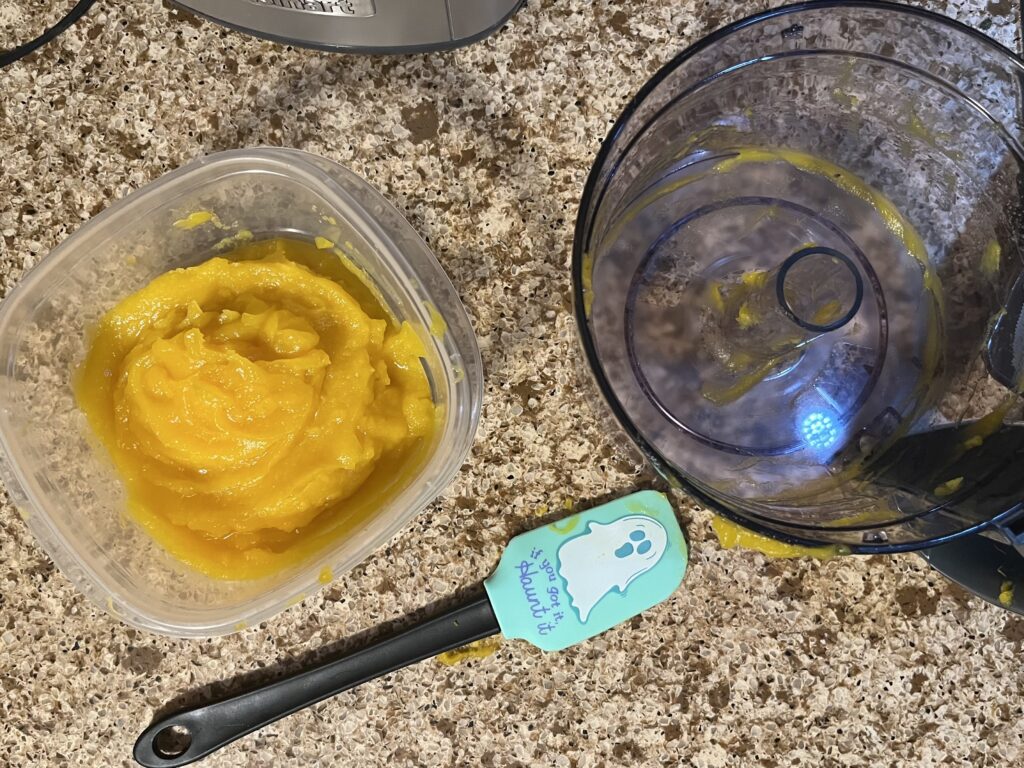 Pumpkin puree in a plastic container, next to an empty food processor and a spatula that has a ghost cartoon on it and the phrase, "If you've got it, haunt it"
