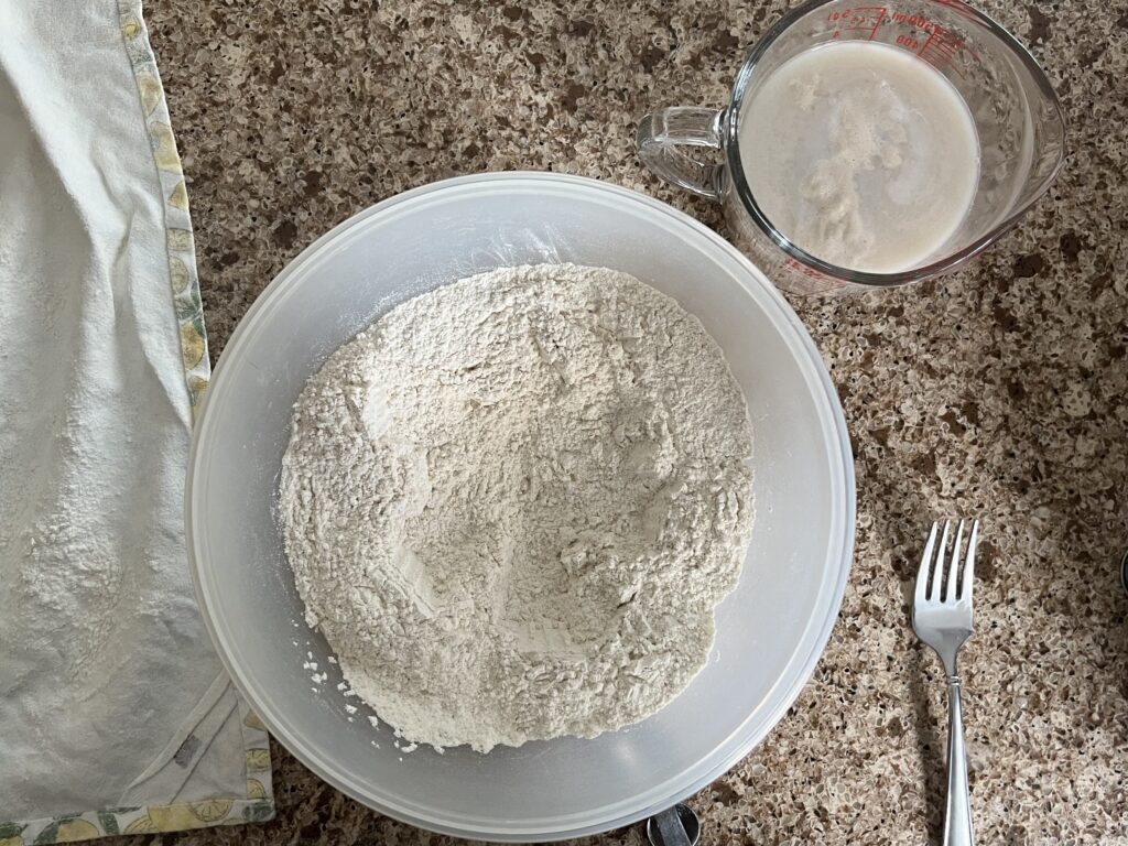 A bowl contains flours and salt, already mixed, and a measuring cup nearby contains water and yeast, which is foaming.
