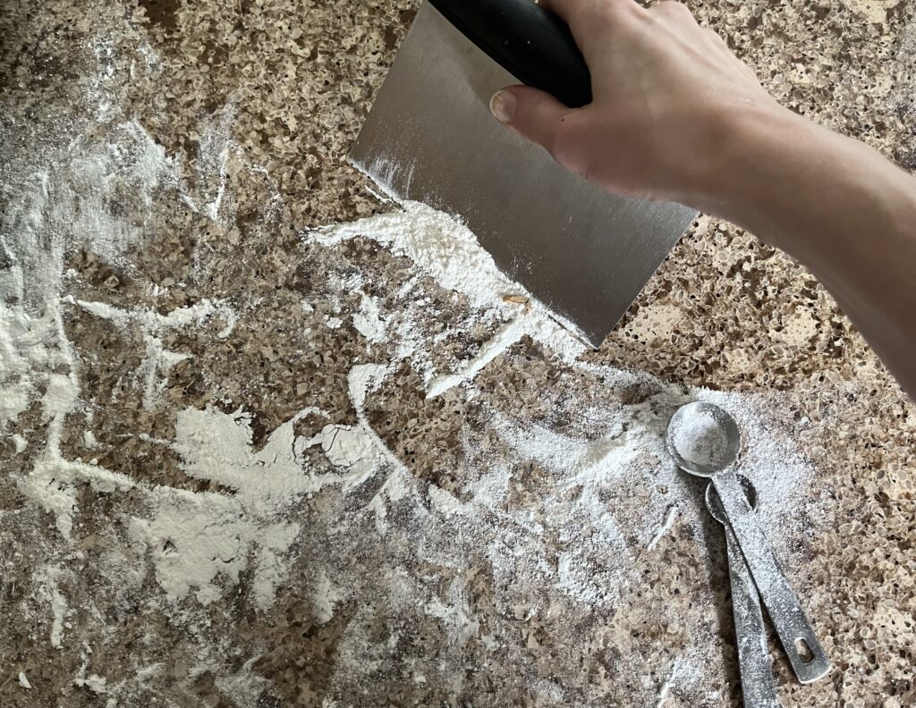 A close-up of someone using a bench scraper to clean flour off a counter top.