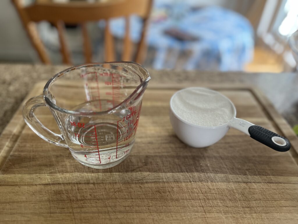 A liquid measuring cup containing one cup of water sits next to a dry measuring cup containing one cup of granulated sugar.