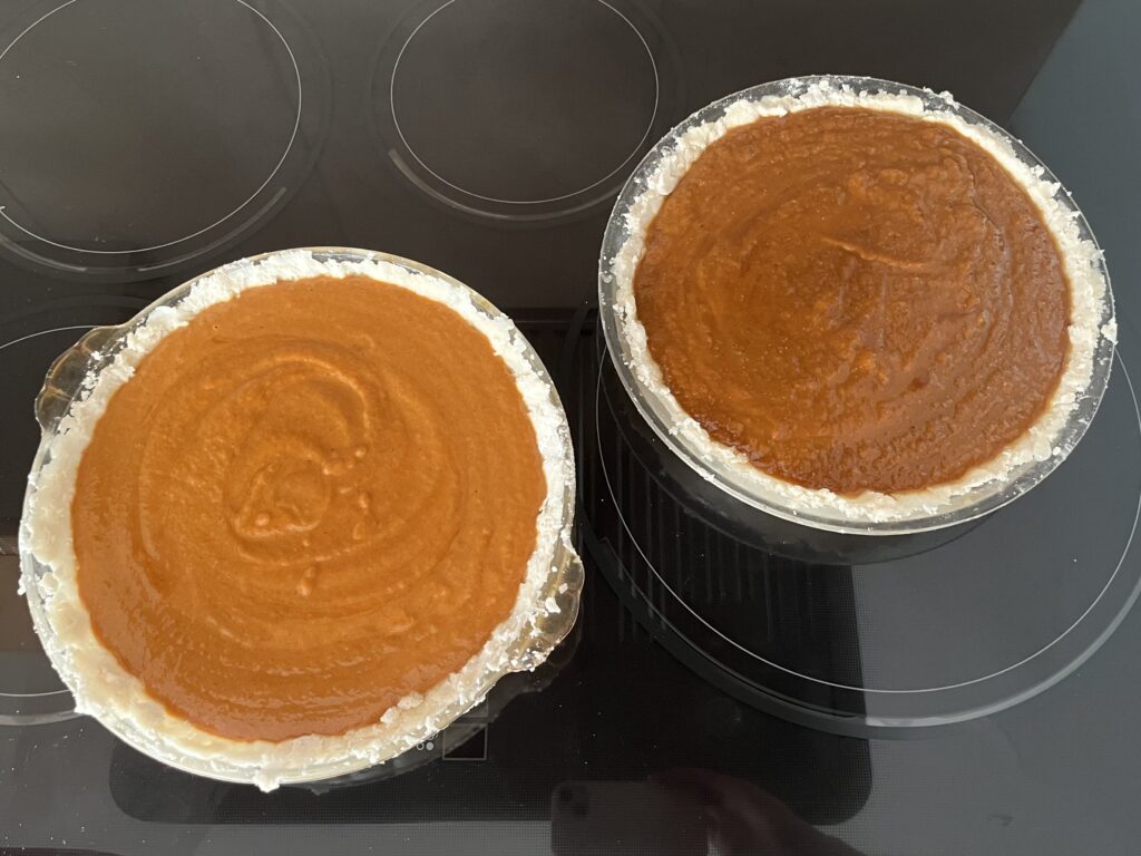 Two unbaked pumpkin pies sit next to each other on a stovetop. The one on the left (traditional) is a lighter orange and the one on the right (vegan) is a darker, golden brown.