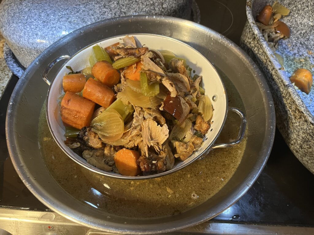 A metal colander sits in a larger metal bowl. The colander contains cooked vegetables and bits of carcass, and soup stock has drained into the bowl.