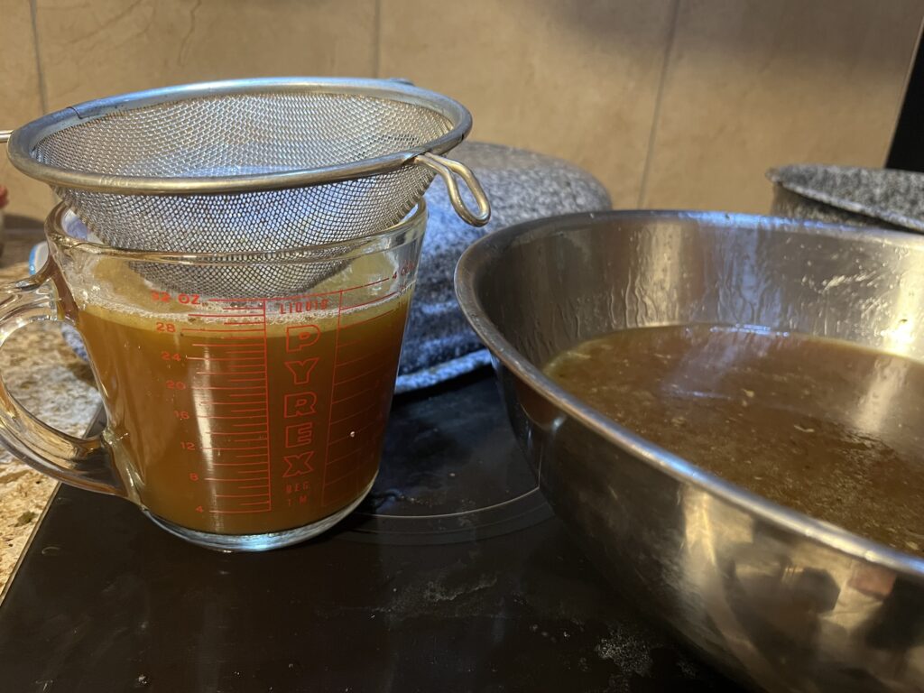 A fine mesh sieve sits atop a liquid measuring cup, which is full of soup stock. Beside it, a large metal bowl contains more stock.