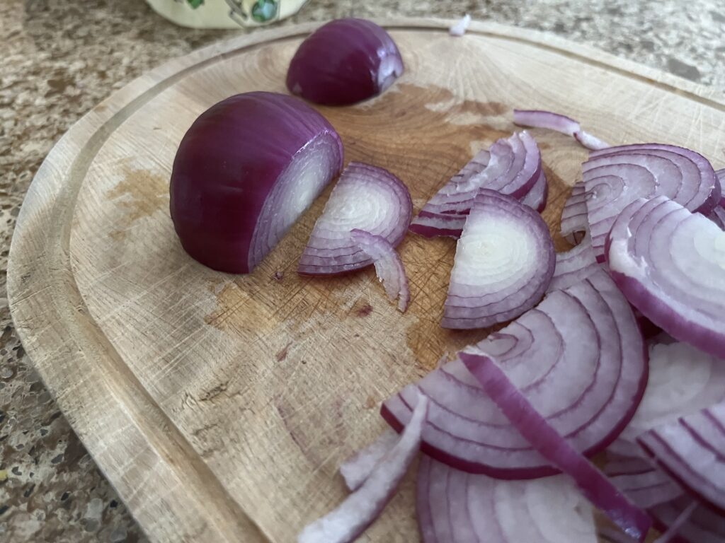 A red onion sits on a cutting board. Half of it has been thinly sliced.
