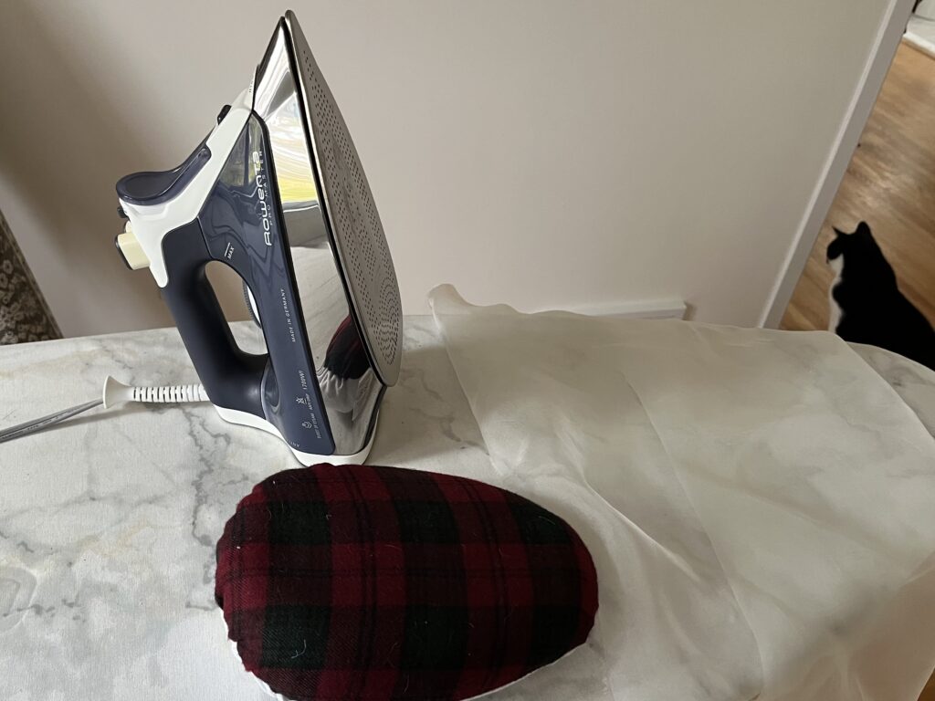 Amy's Rowenta iron on an ironing board with a silk organza pressing cloth and a tailor's ham. A black and white cat is in the background.