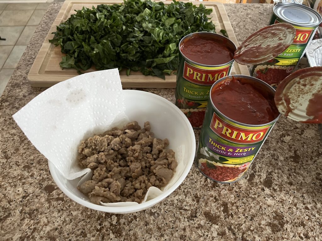Cooked sausage meat in a glass bowl, with opened cans of pasta sauce, and a cutting board covered in chopped spinach in the background.