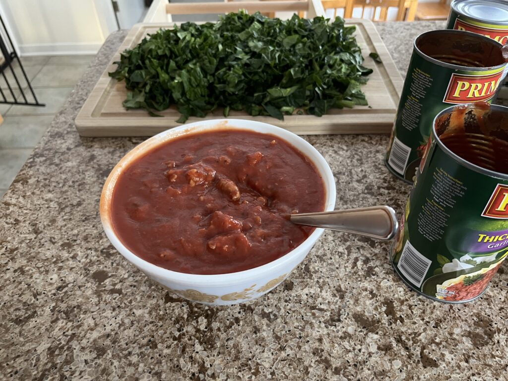 A glass bowl full to the brim with tomato sauce mixed with sausage meat. Chopped spinach is in the background on a cutting board.