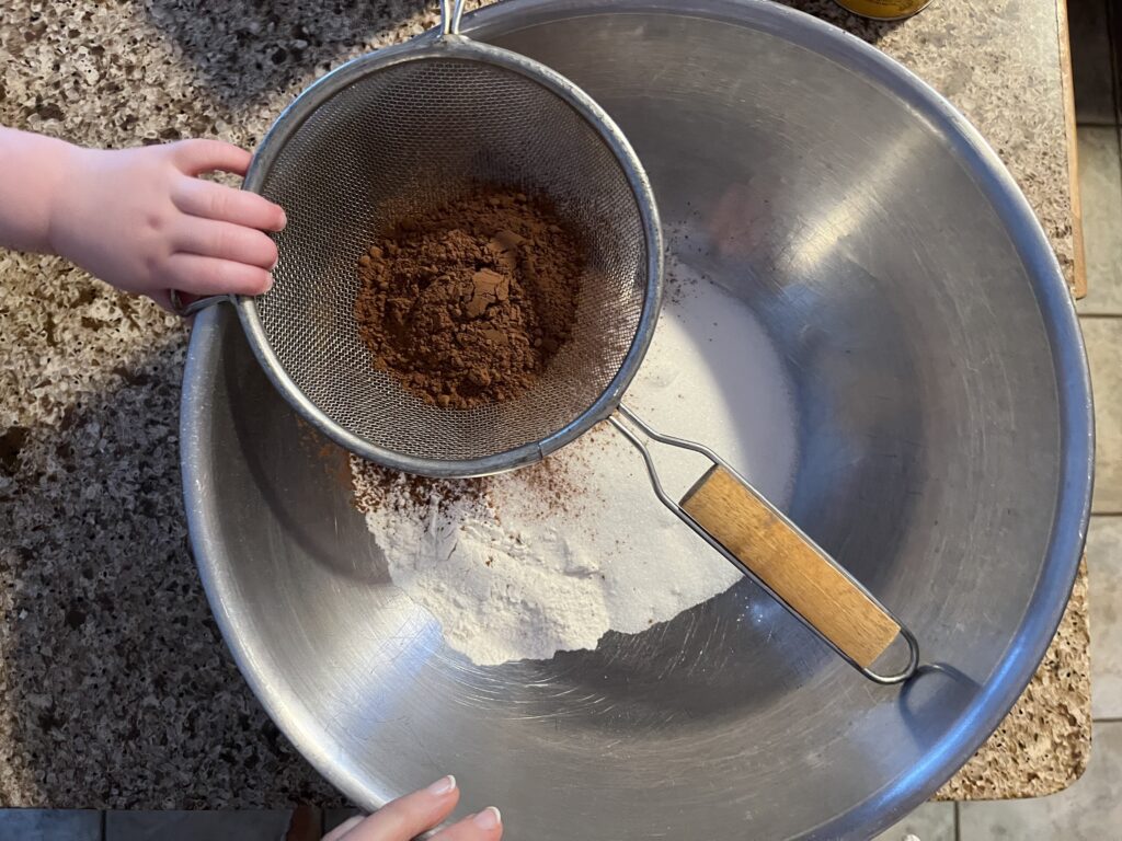 A toddler's hand holds a sieve filled with cocoa over a large metal bowl containing flour and sugar.